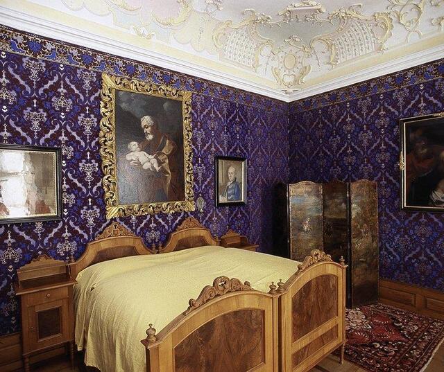 Camera da letto storica nel Museo provinciale altoatesino della caccia e della pesca, con letto in legno, carta da parati decorata e dipinti.