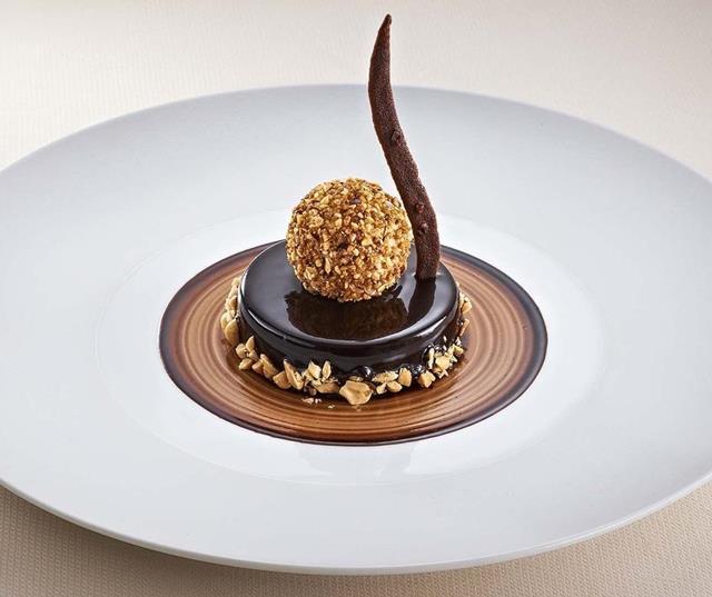 Dessert con glassa al cioccolato, croccante di noci e decorazione al cioccolato su piatto bianco.