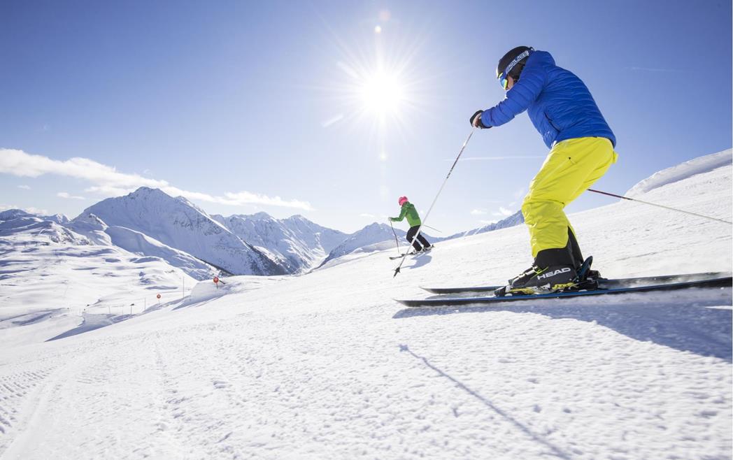 Due sciatori su una pista da sci soleggiata davanti a montagne innevate.