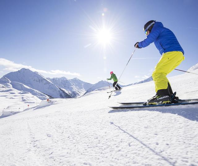 Zwei Skifahrer auf sonniger Skipiste vor verschneiten Bergen.