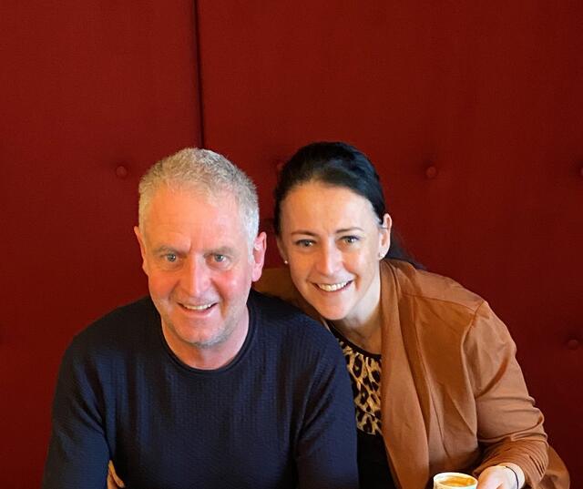 Evelyn e Werner sono seduti sorridenti su una panca rossa e tengono ciascuno una tazza di caffè in mano.