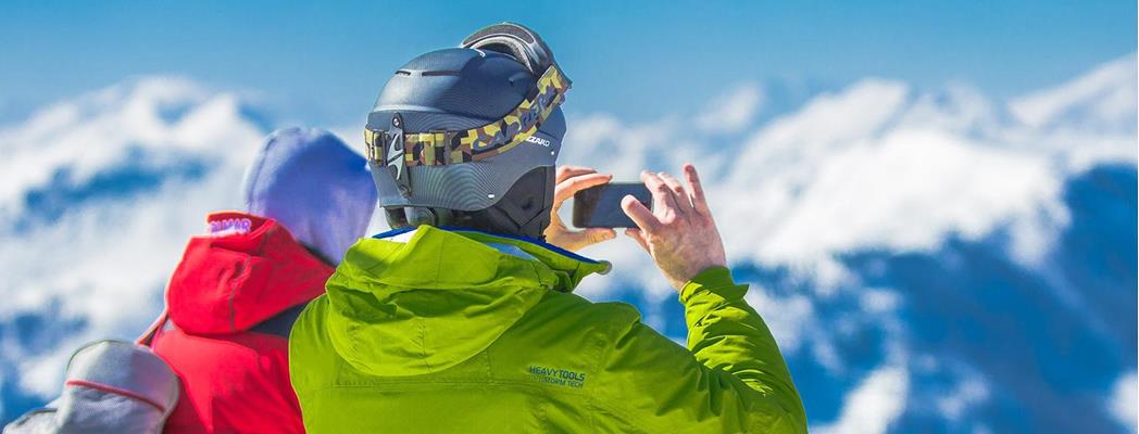 Paar in Winterkleidung blickt auf verschneite Berge; eine Person fotografiert das Winterpanorama.