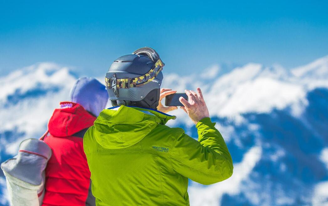 Paar in Winterkleidung blickt auf verschneite Berge; eine Person fotografiert das Winterpanorama.
