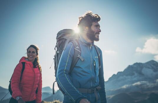 Zwei Wanderer mit Rucksäcken stehen bei Sonnenschein in alpiner Berglandschaft.