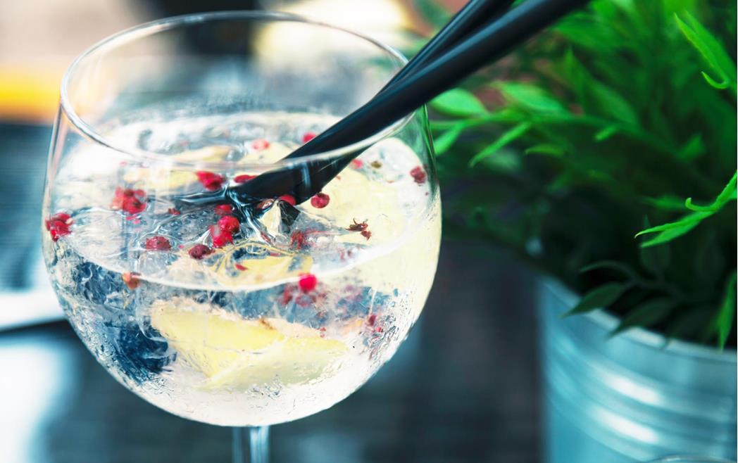 Primo piano di un cocktail in un bicchiere con cubetti di ghiaccio, una fetta di limone, bacche di pepe rosa e una cannuccia nera.