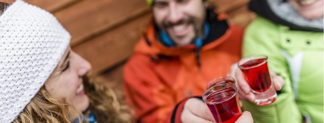 Tre persone in abbigliamento invernale brindano con bevande rosse davanti a una parete in legno.