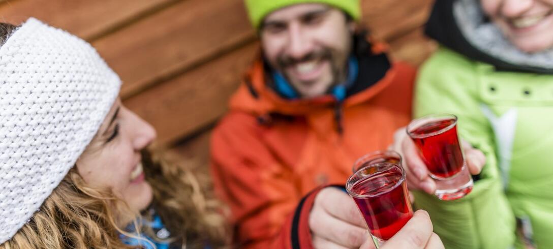 Tre persone in abbigliamento invernale brindano con bevande rosse davanti a una parete in legno.