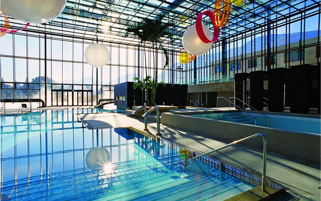 Therme Meran - Innenpool mit Glasdach, großen Fensterfronten und Liegen am Beckenrand.