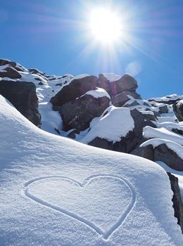 Herz im Schnee vor verschneiter Felslandschaft bei Sonnenschein.