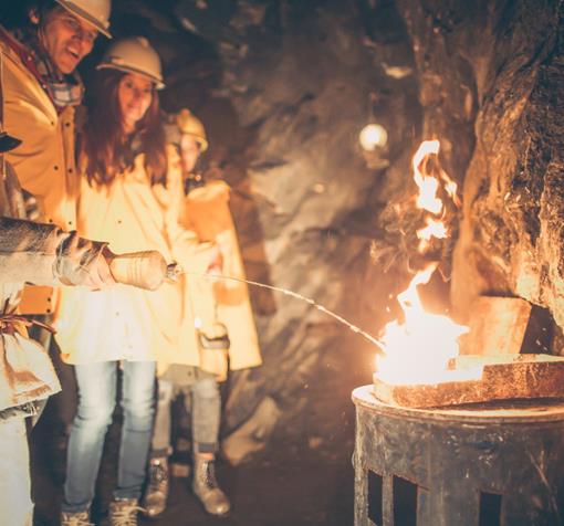 Kinder und Erwachsene mit Helmen stehen in einem Bergwerksstollen und betrachten ein Feuer an der Felswand.