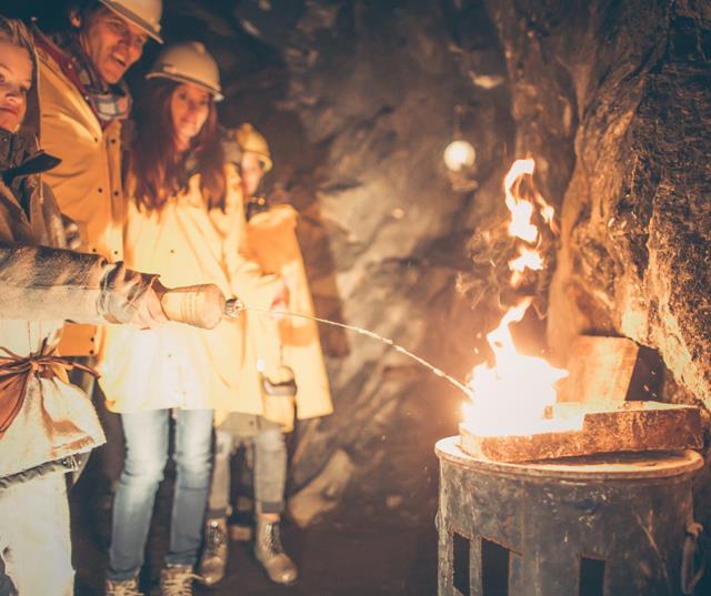 Children and adults wearing helmets stand in a mine tunnel and look at a fire on the rock wall.