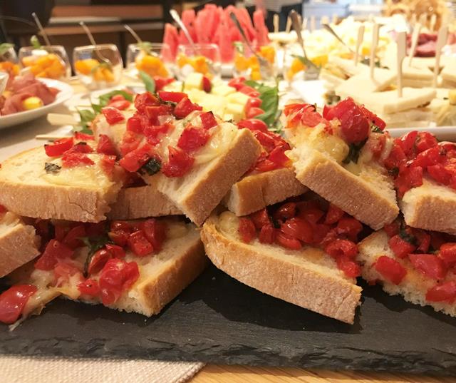 Diversi stuzzichini; in primo piano, fette di pane farcite con pomodori, erbe aromatiche e altri ingredienti, disposte su un vassoio.